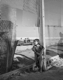 Photo of a woman sitting against a mesh wall, behind her a car with men