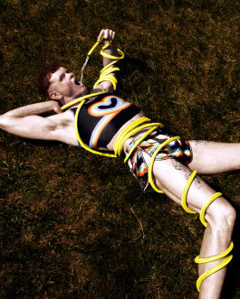 A man lies on the grass, wrapped in a hose from the end of which he pours water into his mouth, dressed in a swimsuit from the Effenberger Couture rainbow collection