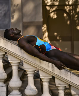 A black man lies on a stone railing outside in a bathing suit from Effenberger Couture x Timo Gerber