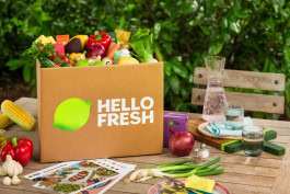 There is a HelloFresh box full of vegetables on the table, next to it are recipe cards, vegetables and two glasses of water next to the bottle 