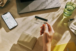 A person holds a bank card in his hand, against the background of a table on which lies a wallet, a phone with an open N26 app, a laptop and other things. 