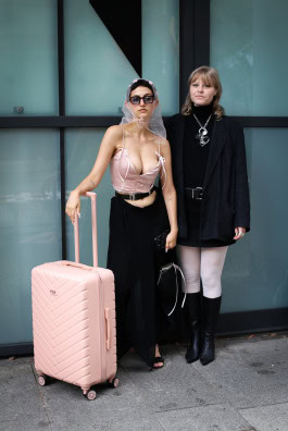 Two women are posing: on the left she is dressed in a long black skirt, a pink corset, with a pink suitcase standing next to her, and on the right she is dressed in a black short outfit and black boots