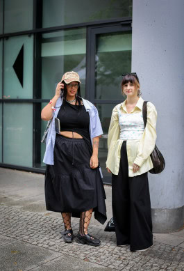 Two women posing: on the left wearing a long black skirt with mesh tights and a black top, and on the right wearing black loose pants, a yellow shirt and a yellow corset