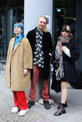 Three people posing: a girl with blue hair dressed in red pants and a beige coat, a guy in red jeans and a black printed shirt and a girl in a black short dress with a printed scarf