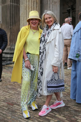 Two women posing: on the left she is wearing printed light pants, a yellow T-shirt, a yellow coat and a yellow bucket hat, on the right she is wearing a white trench coat and pink sneakers