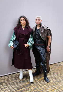 A woman and a man are posing: the woman is wearing a brown coat and white boots, the man is wearing a black leather suit and a leopard print raincoat