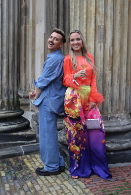 A woman and a man are posing: a man in a denim suit and a woman in multi-colored loose pants and an orange sweater