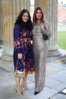 Two women posing: on the left in a blue dress with a floral print, on the right in a silver shiny tight long dress