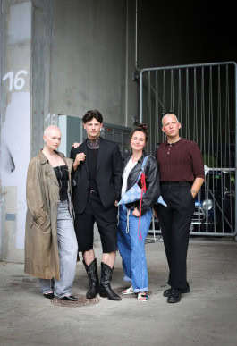 Four people posing: a woman with short shaved white hair and a long raincoat, a man in a black suit with shorts, a woman in light jeans and a man in black trousers and a brown blouse