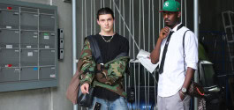 Two guys are standing next to each other, one in a khaki bomber jacket, the other in a white shirt and green cap.