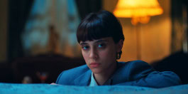 Still from the film Spirit of Ecstasy where a woman looks at the camera with her hand on the edge of a bed