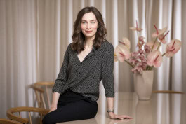 Alexandra Laubrinus half-sitting on a table, with a large vase of flowers on the table in the background