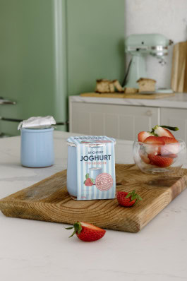 In the kitchen there is a board on which there is a package of yogurt and cut strawberries