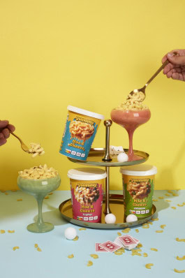 A stand on which there are three packages of Mac and Cheese, as well as glasses where hands are poured with Mac and Cheese