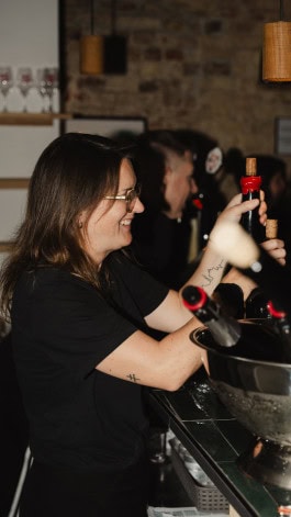 A female bartender smiles and holds bottles of wine behind the bar at Blanc de Fck bar in Berlin