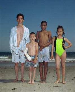 photo of three little boys and one little girl in bathing suits on the beach with the sea in the background, one of the boys has a plaster cast on his arm