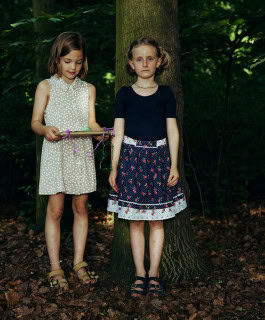 photo of two little girls in a garden, one standing with her back against a tree while the other looks at a game board in her hands