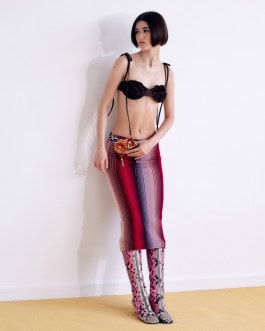 A girl with a dark bob stands against the wall wearing a black bra with flowers on the cups and a medium pink-purple skirt with vertical stripes and a braided belt with flowers on the buckle, on her feet she has purple-gray boots with a zebra print