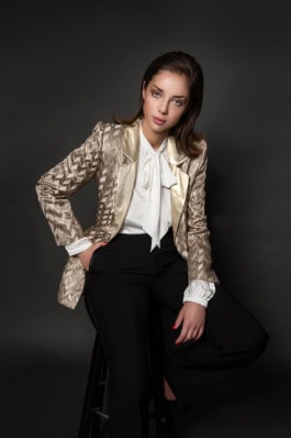 Girl with slicked back hair sitting on chair in suit with black pants, beige shiny jacket and white shirt