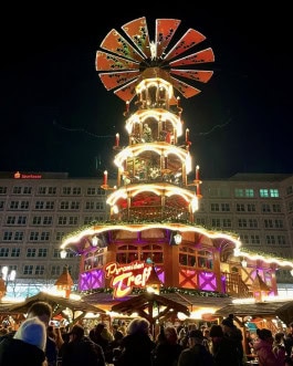Berlin Christmas Market, crowds stroll between decorated Christmas stalls, with a large pyramid of Christmas decorations towering above them 