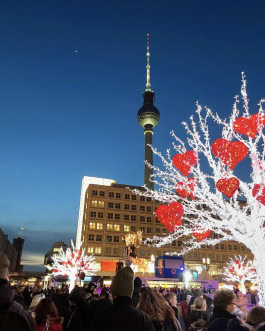 Berlin Christmas Market, crowds stroll past large white trees festooned with flowers and the Berlin TV Tower 