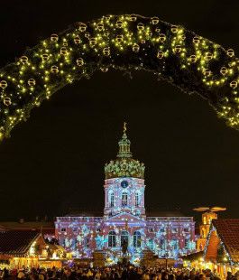 Berlin Christmas Market, a crowd of people walks in front of a building with projections of snowflakes on it and an arch of decorated fir branches 