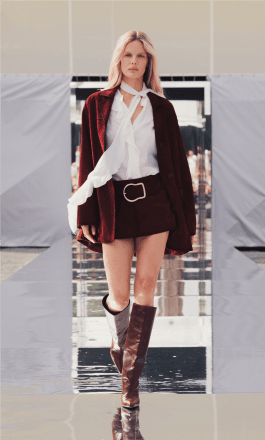 A girl with blonde hair walks the catwalk in burgundy high-heeled boots and a burgundy suit consisting of a short skirt with a wide belt, a jacket, a white shirt and a white thin scarf 
