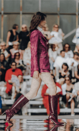 A girl with dark hair walks the catwalk in plum heeled boots and a plum suit consisting of a short skirt, a jacket and a white shirt 