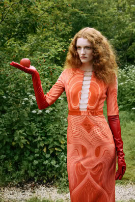 Red-haired curly girl in nature in an orange dress with a cutout along the chest and orange gloves holds an apple in her palm 