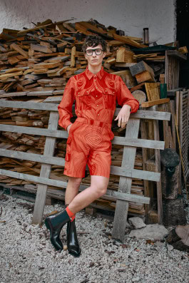 A man in glasses, boots and an orange jumpsuit with shorts stands leaning on a wooden crossbar fencing firewood 