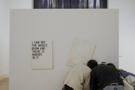 Photo of the Rirkrit Tiravanija exhibition, where three people are leaning over a white stand and looking at one point, next to them hangs a poster with the inscription