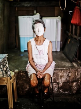 Photo by Kiri Dalena of an elderly Asian woman sitting on a chair with her face covered in white paint 