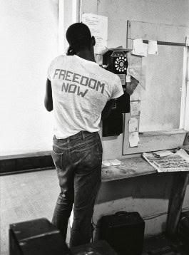 Camera Work exhibition, black and white photo of a black man with his back turned and talking on a pay phone in a room, the inscription on his T-shirt