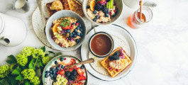 Table with a healthy breakfast: two bowls of oatmeal with fruits and berries, a bowl of yogurt with fruits and berries, a waffle with honey and blackberries, a cup of coffee, a teapot, a pitcher of milk, a jar of honey and a bouquet of flowers