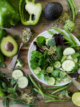 Chopped green vegetables and herbs in a plate and on the table: cucumbers, spinach, avocado, green peppers, peas, broccoli, mint, wheat sprouts, parsley