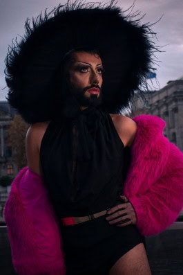 The drag queen poses in a black bodysuit, a pink short fur coat and a wide black fur hat 